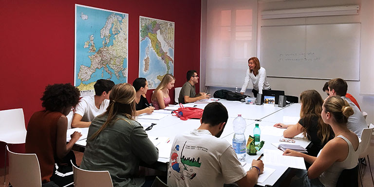 Students seated around a table listen to a speaker standing at the head of the table.