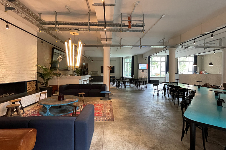 A wide, open space, showing a seating corner with couches on the left, a long, turquoise table on the right and a few other smaller tables in the back of the room