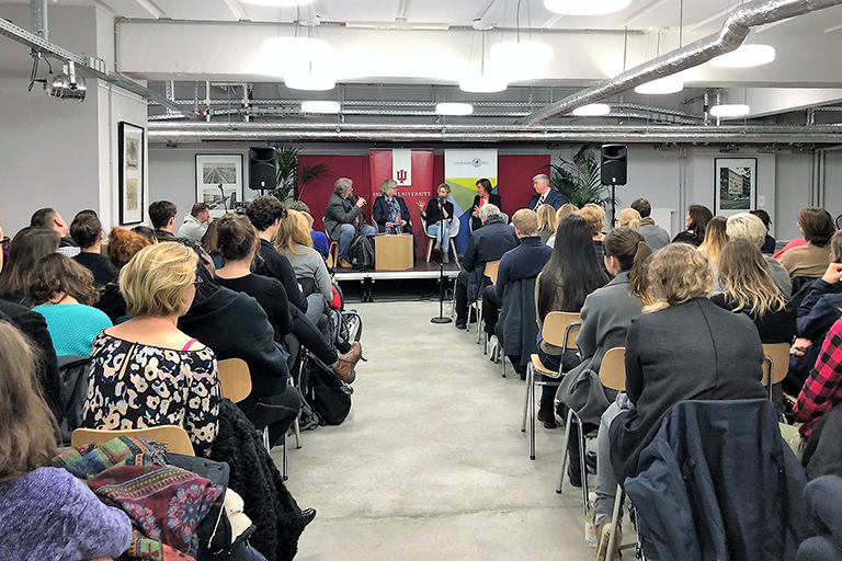 A public event with a panel on a small stage and two rows packed with a lot of people.
